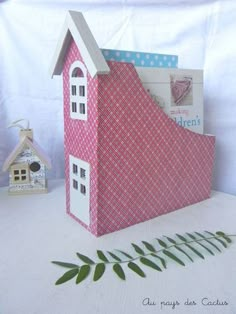 a small pink house sitting on top of a table next to a green plant and white wall