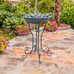 a bird bath sitting on top of a metal stand next to some flowers and trees