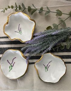 three white dishes with purple flowers and greenery in them on a striped tablecloth