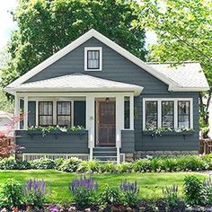 a small gray house with white trim and flowers in the front yard, on a sunny day