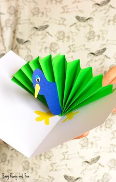 a hand holding a paper bird with green wings and blue beak, on top of a white card