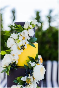 a yellow and black wedding cake with white flowers on the top, surrounded by greenery