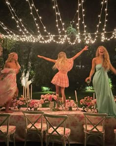 three women in dresses standing on a table with lights strung above them and flowers all around