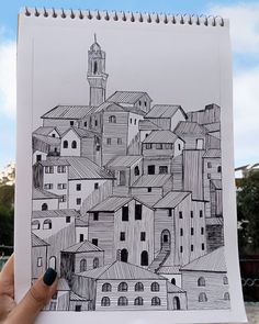 a hand holding up a drawing of a city with buildings and a clock tower in the background