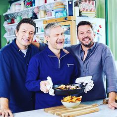 three men standing in front of a table with food on it and one holding a frying pan