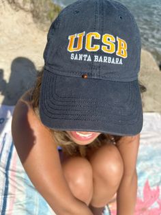 a woman sitting on top of a towel next to the ocean wearing a baseball cap