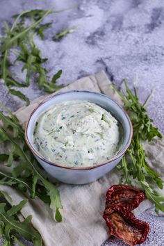 a blue bowl filled with white sauce next to some green leaves and red peppers on a cloth