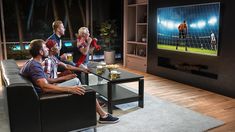 four people sitting around a table watching tv with drinks in front of them and one man holding a beer