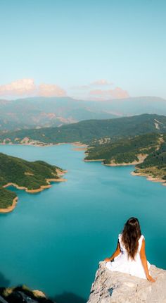 a woman sitting on the edge of a cliff overlooking a lake with mountains in the background