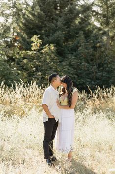 an engaged couple kissing in the middle of a field with tall grass and trees behind them