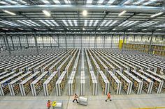 two workers are walking in front of rows of shelving units inside a warehouse with overhead lighting