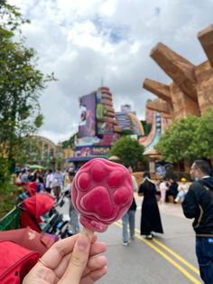 a person is holding up a pink ice lollypop in front of an amusement park