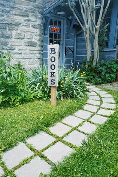 a sign that is in the grass near a house with flowers and plants around it