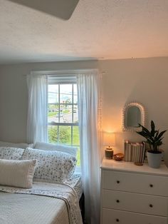 a bedroom with a bed, dresser and window in the corner that has white curtains