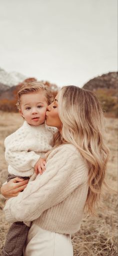 a woman holding a baby in her arms and kissing the child's face with mountains in the background