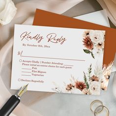 an orange and white floral wedding response card with gold rings on the table next to it