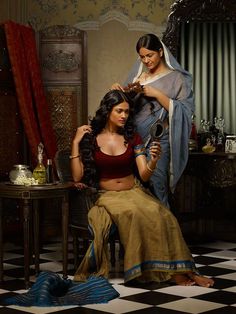 two women are sitting in a room with black and white checkered flooring, one is cutting the other's hair