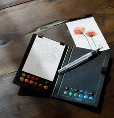 an artist's notebook with watercolors and a pen on the table next to it