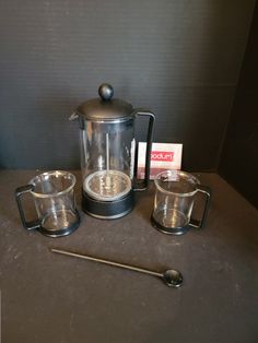 a coffee pot and two cups on a table with a spoon next to it,