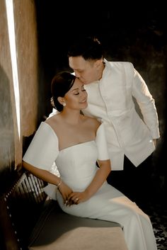 a man and woman sitting next to each other in front of a wall with their arms around each other