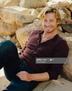 a young man sitting on the ground next to rocks and smiling at the camera with his hands in his pockets