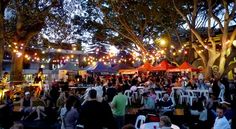 a group of people sitting around tables under trees with lights strung from the branches over them