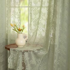 a white vase with flowers on a table in front of a curtained room window