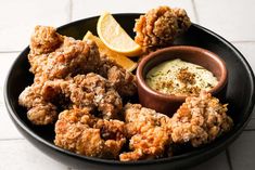 a black plate topped with fried food next to a bowl of dip and lemon wedges