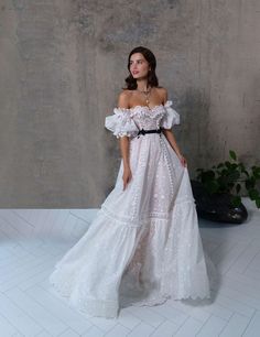 a woman wearing a white dress with ruffles and a black belt standing in front of a concrete wall