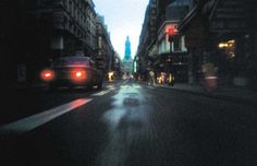 a car driving down a street at night with buildings in the background and people walking on the sidewalk