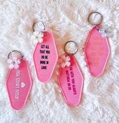 three pink keychains with words on them sitting on a white blanket, one has a flower and the other has a ring