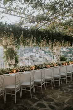 an outdoor wedding setup with white chairs and greenery on the wall, overlooking a cityscape