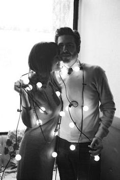 a man and woman standing next to each other in front of a window covered with christmas lights