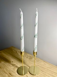 two white candles sitting on top of a wooden table next to each other with gold bases