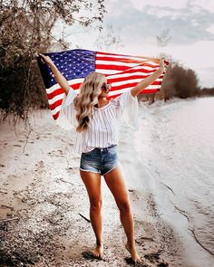 a woman holding an american flag in her hands on the shore of a lake or beach