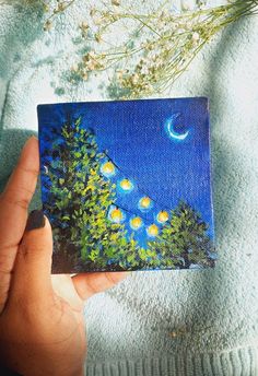 a hand holding up a small painting on a blue surface with trees and the moon in the sky