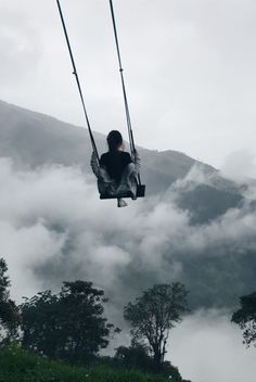 a person sitting on a swing in the air with clouds and trees behind them,