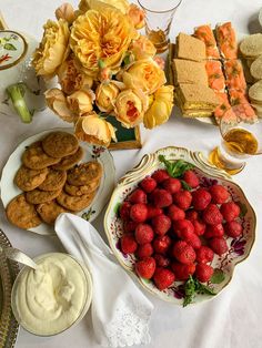 a table topped with plates filled with desserts and fruit next to flowers on top of a white table cloth