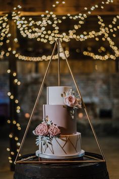 a white and pink wedding cake on top of a wooden barrel with lights in the background