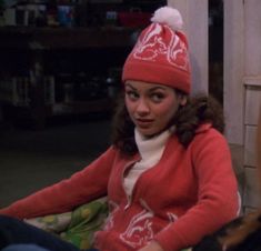 a woman sitting on the floor wearing a red sweater and a white pom - pom hat