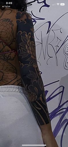 the back of a woman's body with tattoos on her arm and shoulder, in front of a graffiti wall