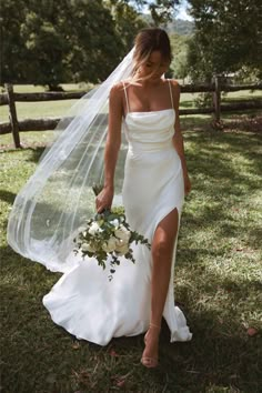 a woman in a white wedding dress and veil walking through the grass with her bouquet