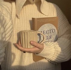 a woman holding a book and coffee mug in her hands while wearing a white sweater