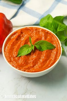 a white bowl filled with tomato sauce next to some fresh basil leaves and red peppers