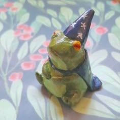 a small frog figurine with a hat on it's head sitting on a flowered surface