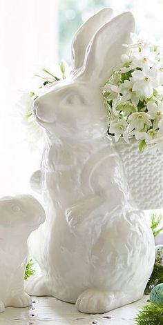 two white ceramic rabbits sitting next to each other on a table with flowers in the background