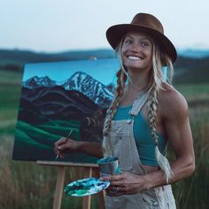 a woman in overalls holding a paintbrush next to an easel with a painting on it