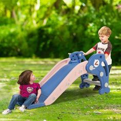 two children playing on a blue and pink slide in the grass with trees in the background