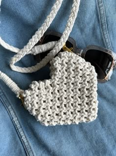 a crocheted purse sitting on top of a pair of blue jean pants with sunglasses