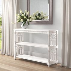 a white console table with flowers on it in front of a mirror and curtained window